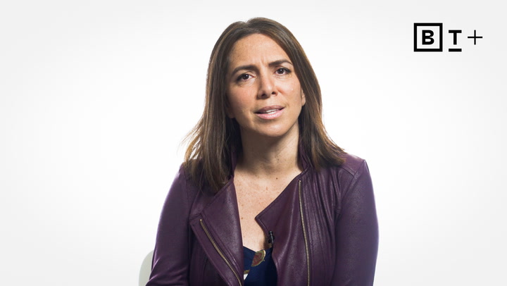 A woman with straight brown hair wears a purple jacket and sits against a plain white background with a "BT+" logo in the top right corner.