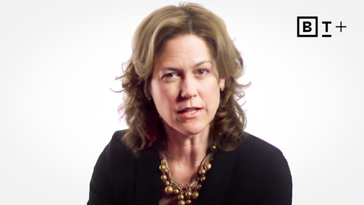 A woman with wavy brown hair wearing a black blazer and beaded necklace speaks in front of a white background with a "B T +" logo in the top right corner.