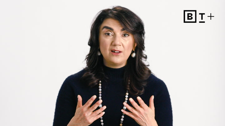 A woman with dark hair and a navy sweater gestures with her hands while speaking in front of a plain white background with a "BT+" logo in the top right corner.