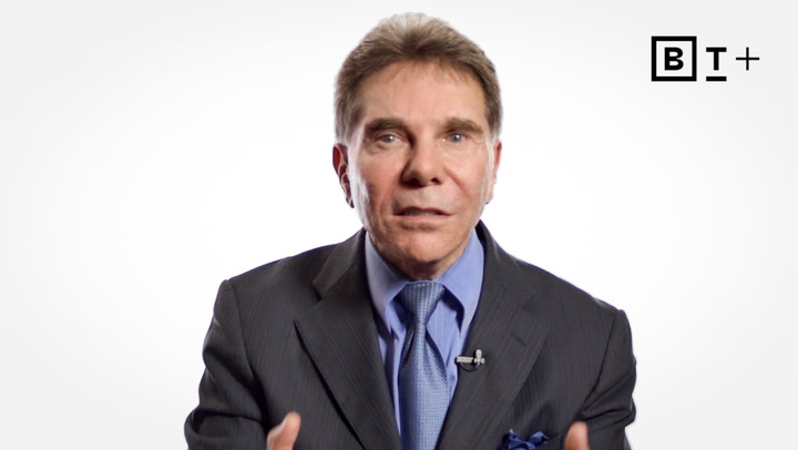 A man in a suit and tie speaks while facing the camera, with a small microphone clipped to his jacket and a simple logo in the top right corner.