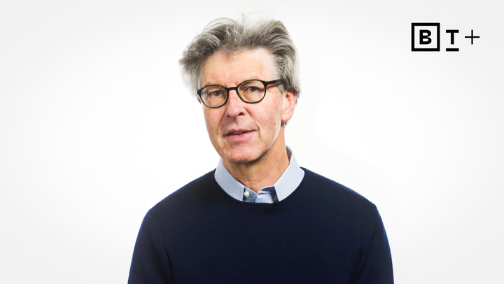 A man with gray hair and glasses, wearing a navy sweater over a collared shirt, stands against a plain white background with a “B T +” logo in the corner.
