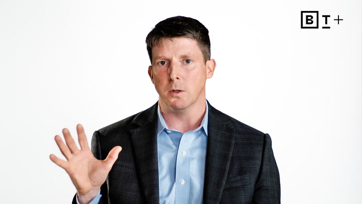 A man in a suit gestures with his right hand raised, standing in front of a plain white background with a "BT+" logo in the top right corner.