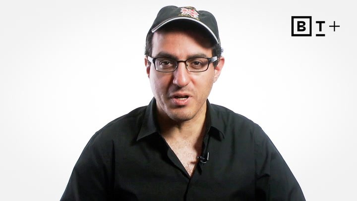 A man wearing glasses, a black shirt, and a black cap is speaking in front of a white background with a "BT+" logo in the top right corner.