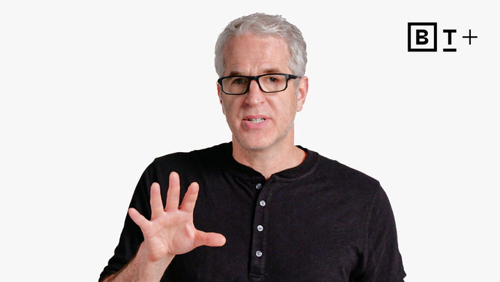 A man with gray hair and glasses in a black henley shirt gestures with his right hand against a plain white background with a "B T +" logo at the top right.