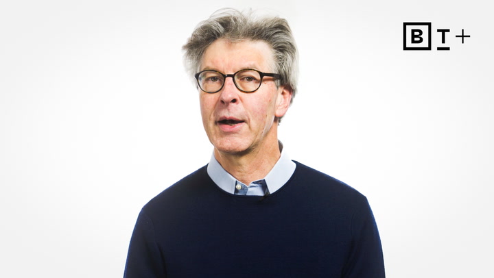 A middle-aged man with gray hair and glasses wearing a navy sweater and light blue shirt stands against a plain white background with a "B T +" logo in the top right corner.