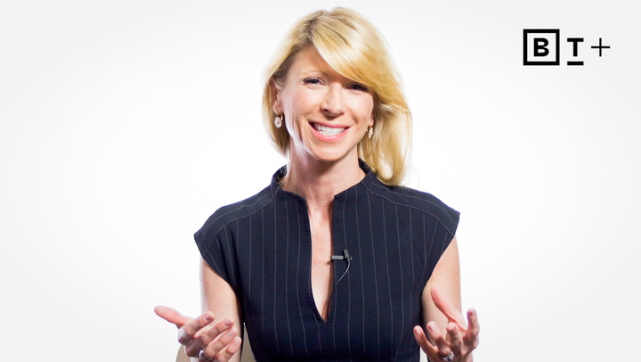 A smiling woman in a dark, short-sleeved dress gestures with her hands while speaking. A microphone is clipped to her collar. The letters "BT+" appear in the top right corner.
