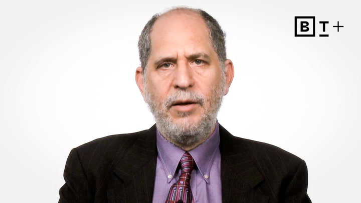 A middle-aged man with a gray beard wearing a suit, purple shirt, and patterned tie is speaking in front of a plain white background with a BT+ logo.