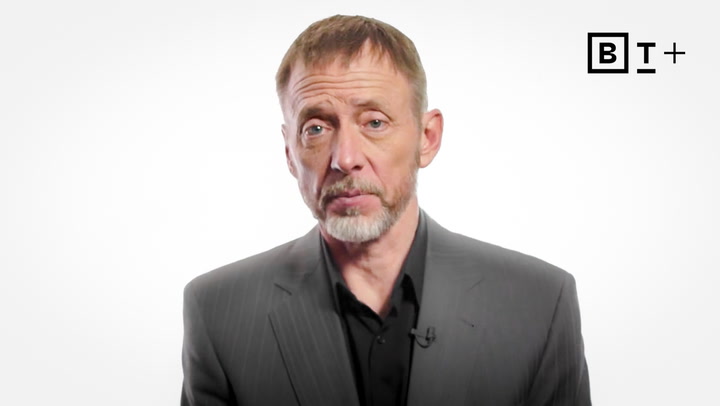 A middle-aged man with short hair and a beard, wearing a dark suit jacket, stands against a plain white background with a "BT+" logo in the top right corner.