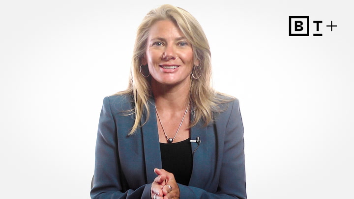 A woman in a gray blazer stands against a white background, smiling at the camera, with a "BT+" logo in the top right corner.