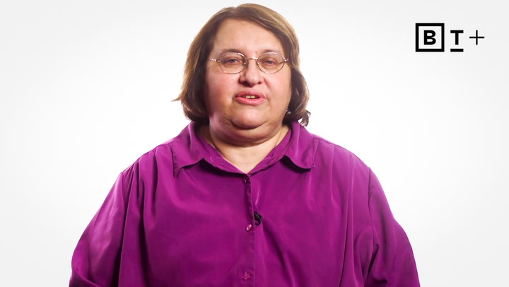A person with short brown hair and glasses wearing a purple shirt poses against a plain white background with a "B T +" logo in the top right corner.