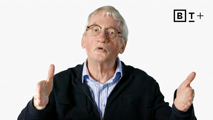 Older man with glasses and gray hair gestures with both hands while speaking; he wears a dark jacket over a striped shirt. "B T +" logo appears in the top right corner.