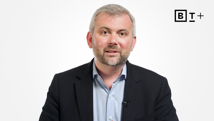 A man in a dark suit jacket and light blue shirt is speaking in front of a white background with a "BT+" logo in the top right corner.