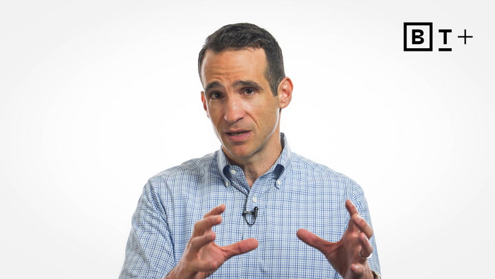 A man in a checkered shirt gestures with his hands while speaking, with a B T+ logo in the top right corner on a plain white background.