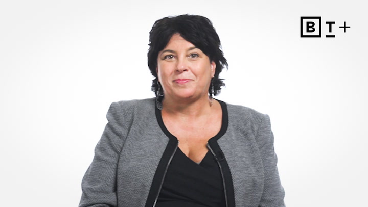 A woman with dark hair, wearing a gray blazer and black top, sits in front of a plain white background with a "B T +" logo in the top right corner.