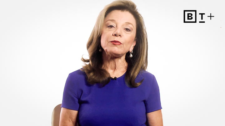 Woman with long brown hair wearing a blue dress sits in front of a plain white background with a "BT+" logo in the upper right corner.