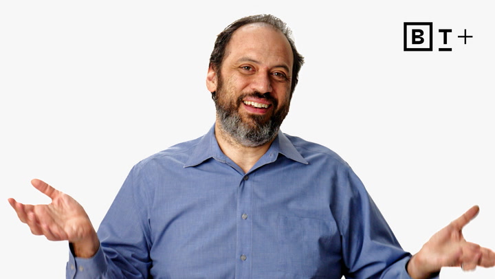 A man with a beard and mustache, wearing a blue button-up shirt, smiles and gestures with his hands raised. The logo "B T +" appears in the top right corner.