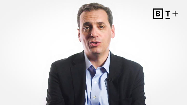 A man in a suit jacket and collared shirt speaks while facing the camera against a plain white background with a "B T +" logo in the top right corner.