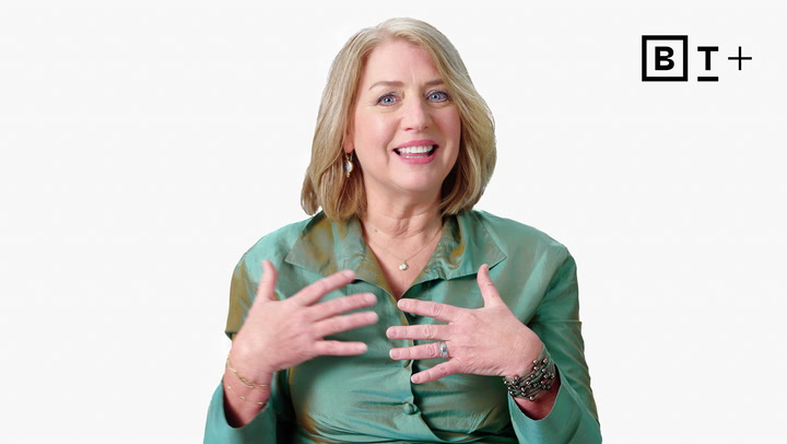 A woman with blonde hair in a green blouse gestures with her hands while speaking, seated in front of a white background with the "B T +" logo.