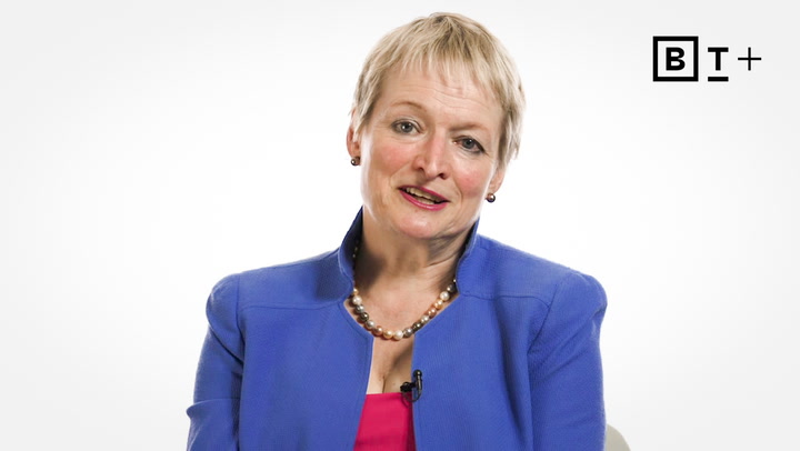 A woman with short blond hair wearing a blue blazer, pink top, and pearl necklace sits in front of a white background with a "BT+" logo in the corner.