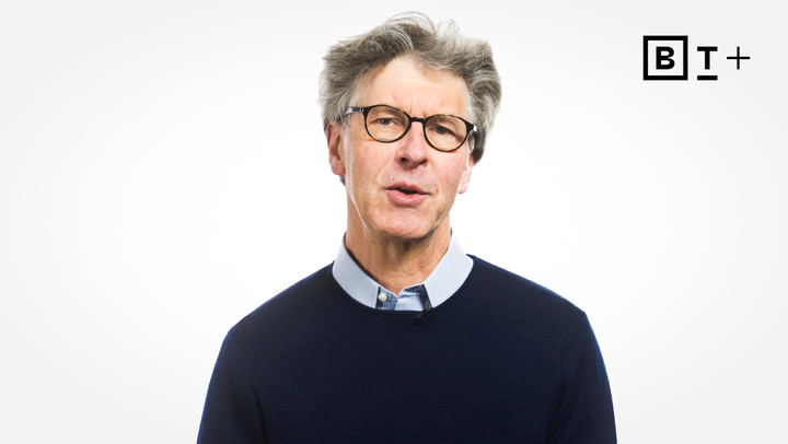 Gray-haired man wearing glasses and a navy sweater stands against a plain white background with a "B T +" logo in the top right corner.