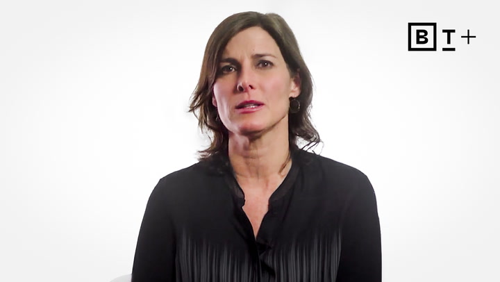A woman with shoulder-length brown hair wearing a black blouse sits against a plain white background with a "B T +" logo in the top right corner.