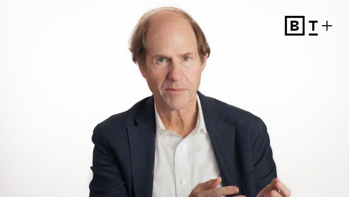 A middle-aged man in a suit jacket and white shirt sits against a plain white background with a "B T +" logo in the top right corner.
