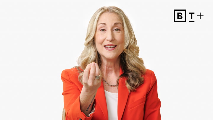 A woman with long blond hair wearing an orange blazer gestures with her right hand while speaking, against a plain white background with a "BT+" logo in the top right corner.