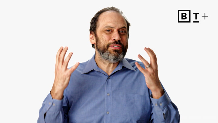 A man with a beard, wearing a blue collared shirt, gestures with both hands while speaking. A "B T +" logo is visible in the top right corner on a plain background.