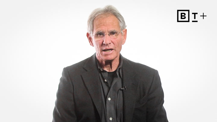 A man with gray hair and glasses wearing a dark suit jacket sits against a plain white background with a "B T +" logo in the top right corner.