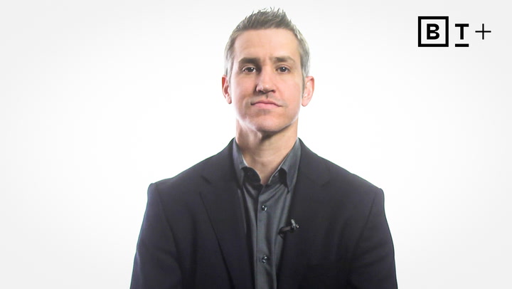 A man in a dark suit jacket and collared shirt sits against a plain white background with a “B T +” logo in the top right corner.