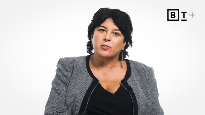 A woman with dark hair wearing a gray blazer and black top speaks while facing the camera; a "BT+" logo is visible in the top right corner.