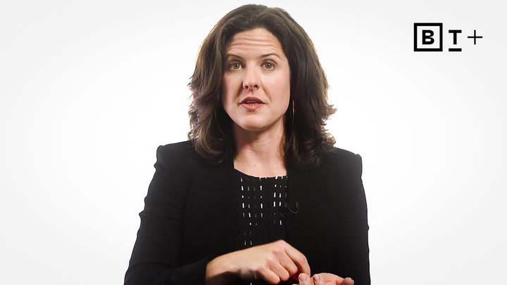 A woman with shoulder-length brown hair, wearing a black blazer, gestures with her hands against a plain white background with a "B T +" logo in the top right corner.