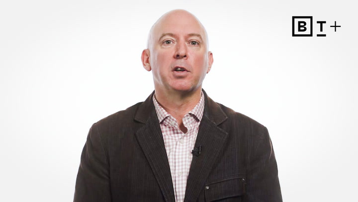 A bald man in a dark blazer and checked shirt speaks in front of a white background with a "B T +" logo in the top right corner.