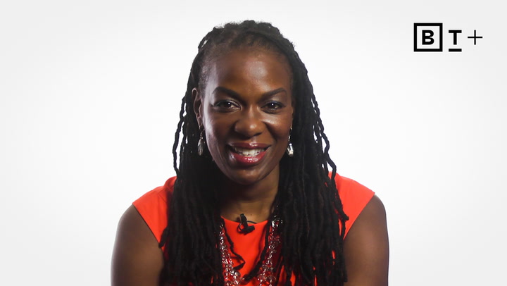 A person with long dreadlocks, wearing a bright orange top and necklace, is smiling at the camera against a plain white background with a "BT+" logo in the corner.