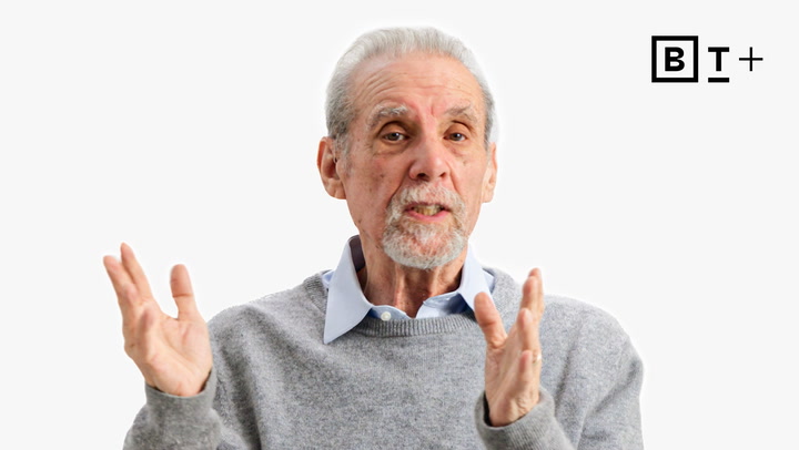 An older man with gray hair and a beard speaks while gesturing with his hands, wearing a gray sweater over a collared shirt; a BT+ logo is in the top right corner.