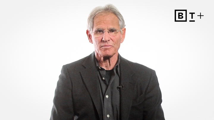 An older man with gray hair and glasses wears a dark suit and shirt, facing the camera against a plain white background with a "BT+" logo in the top right corner.