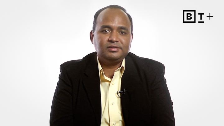 A man in a dark blazer and light shirt sits facing the camera against a plain white background with a "BT+" logo in the top right corner.