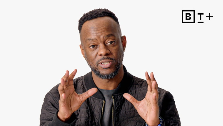 Man in a black jacket gestures with both hands while speaking against a plain white background, with a "B T +" logo in the top right corner.