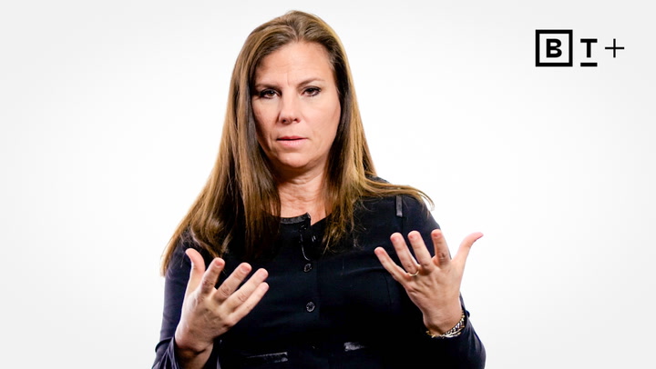 A woman with long brown hair and a black top gestures with both hands while speaking, against a plain white background with a "B T +" logo in the top right corner.