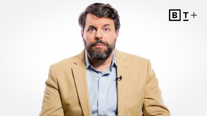 A man with a beard and mustache wearing a tan blazer and blue shirt sits against a plain white background with a "B T +" logo in the top right corner.