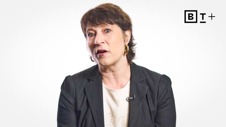 A woman with short brown hair wearing a black blazer and white top sits in front of a white background with a "B T +" logo in the top right corner.