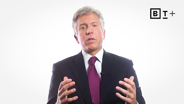 A man in a suit and tie speaks while gesturing with his hands. The background is white with a "B T +" logo in the top right corner.