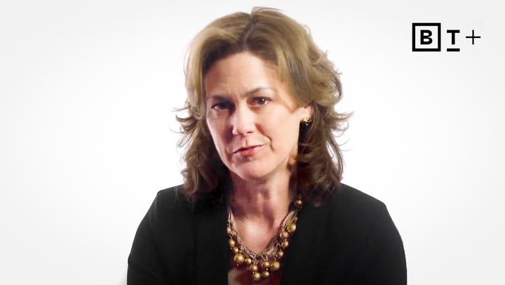 A woman with shoulder-length brown hair wearing a black blazer and a large beaded necklace sits in front of a plain white background. The "BT+" logo is in the top right corner.