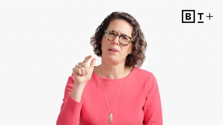 A woman with short curly hair and glasses, wearing a pink top, gestures with her hand pinched, standing in front of a plain white background with a "B T +" logo in the corner.