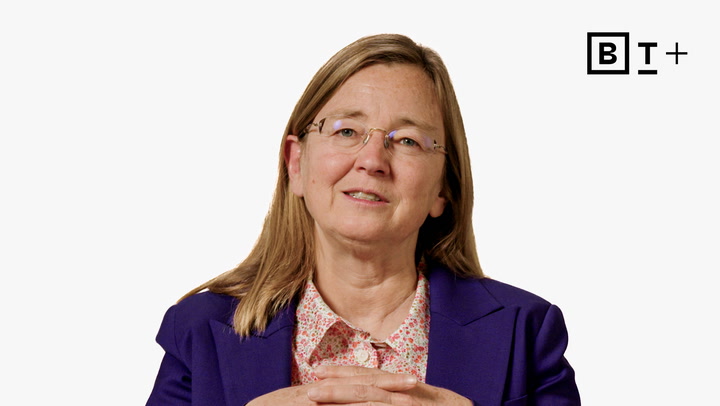 A woman with long light brown hair, wearing glasses, a purple blazer, and a floral shirt, sits with her hands clasped in front of a white background featuring a "B T +" logo.