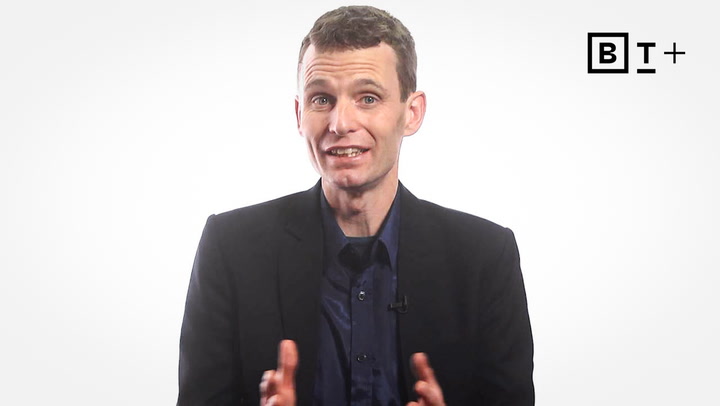 A man in a dark suit and navy shirt gestures with his hands while speaking, standing in front of a plain white background with a BT+ logo in the upper right corner.