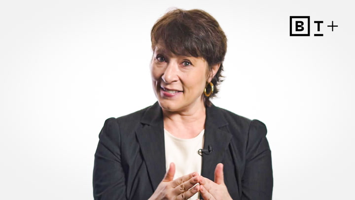 A woman with short dark hair wearing a black blazer and white top gestures with her hands while speaking. A "BT+" logo appears in the top right corner.