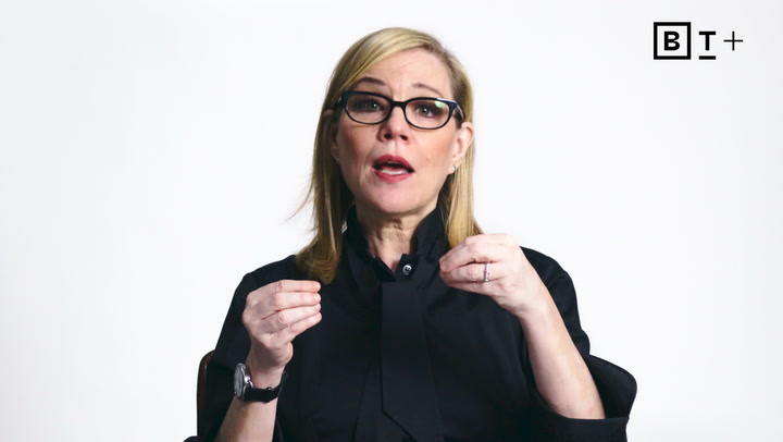 A woman with blonde hair and glasses, dressed in a black top, gestures with her hands while speaking against a plain white background.