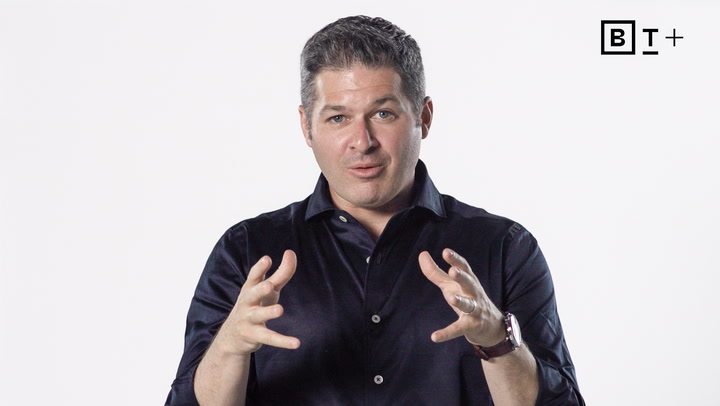A man in a dark shirt gestures with his hands while speaking, against a plain white background with a "BT+" logo in the top right corner.