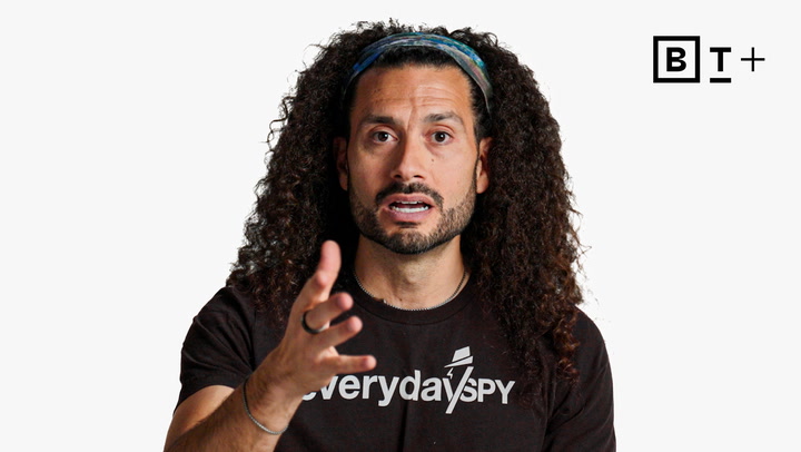 A man with long curly hair wearing a black "Everyday Spy" shirt gestures while speaking, with a "B T +" logo in the top right corner on a white background.